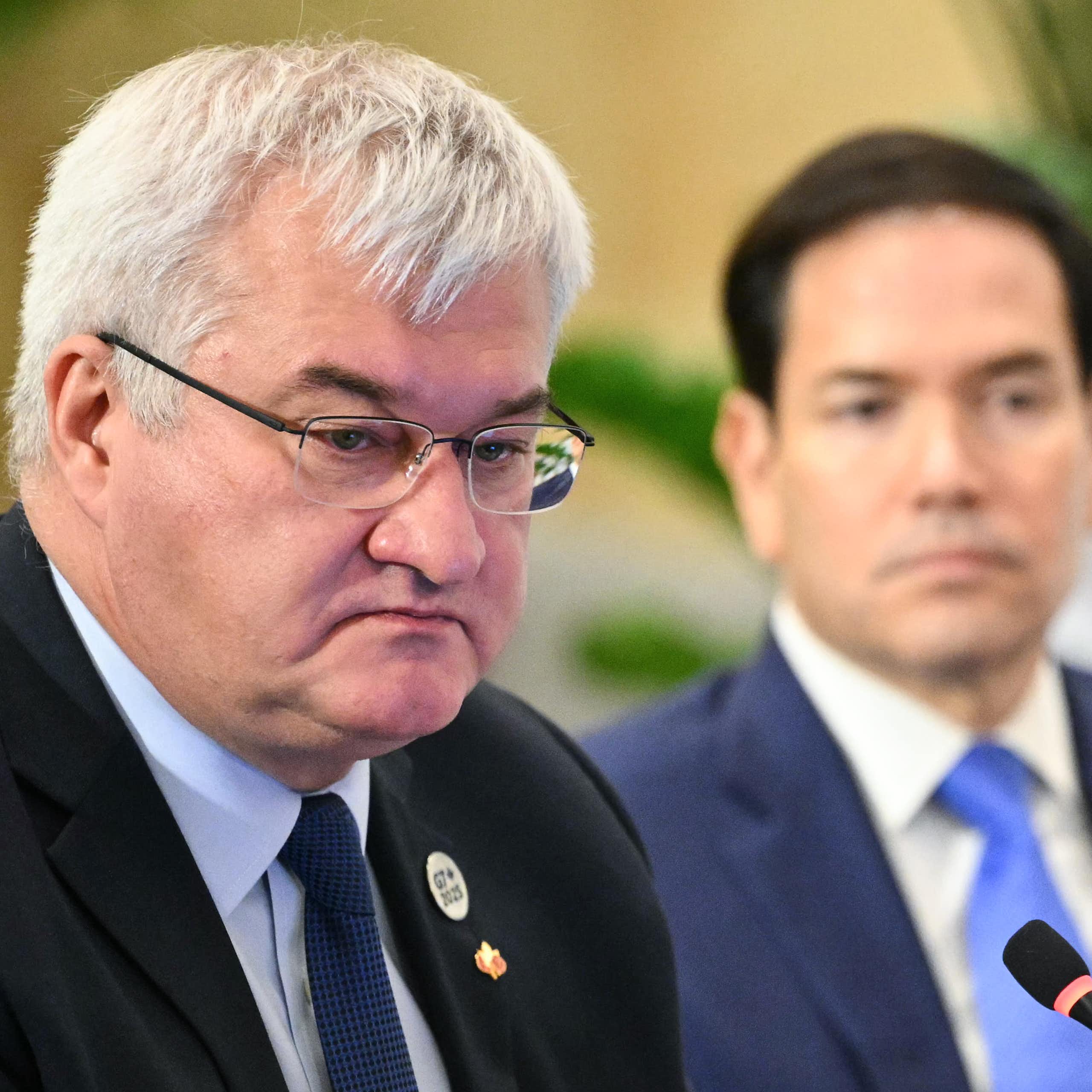 A round-faced grey-haired man wearing glasses with a younger dark-haired man in the background.