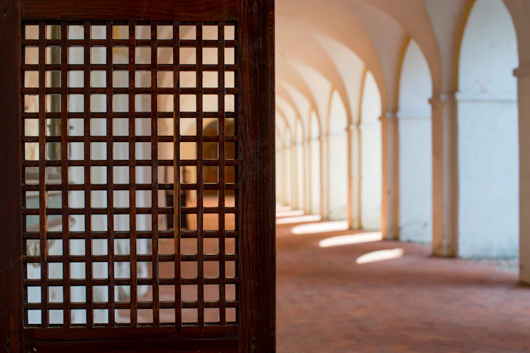 A photograph shows, at left, what appears to be part of a wooden door to a confessional booth in a religious setting, next to a row of arched openings, with sunlight coming out of them, on a walkway.