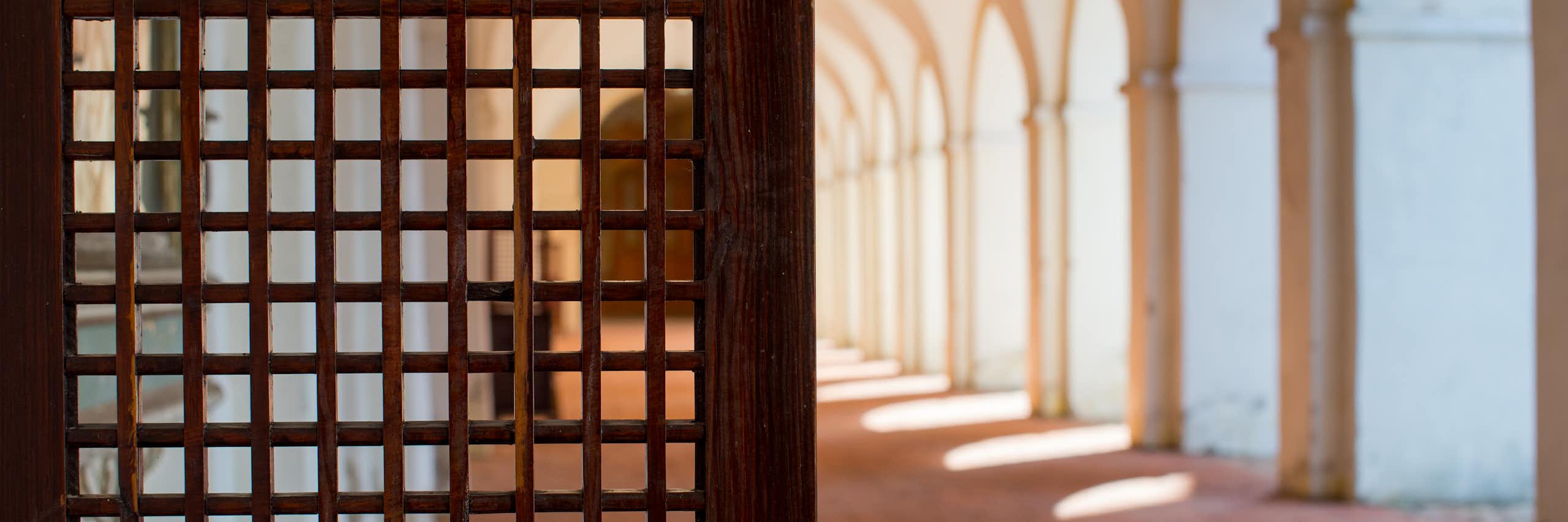 A photograph shows, at left, what appears to be part of a wooden door to a confessional booth in a religious setting, next to a row of arched openings, with sunlight coming out of them, on a walkway.