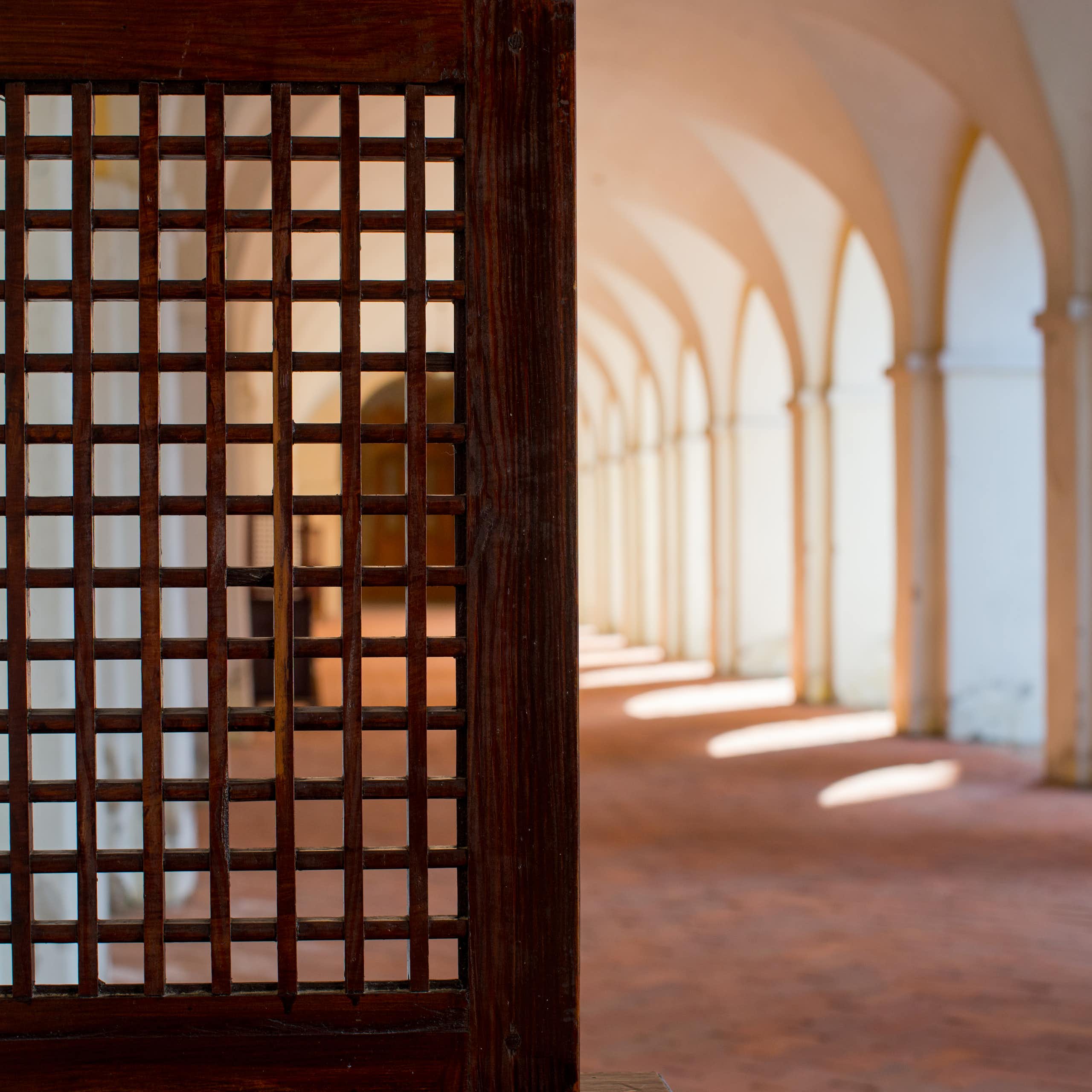 A photograph shows, at left, what appears to be part of a wooden door to a confessional booth in a religious setting, next to a row of arched openings, with sunlight coming out of them, on a walkway.