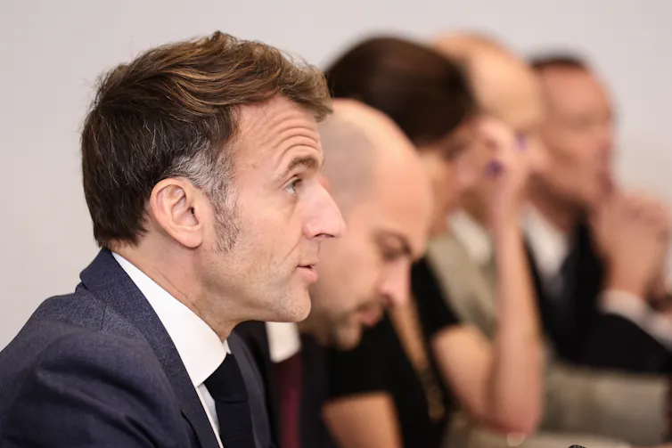 a dark-haired man photographed in profile; other men sit next to him
