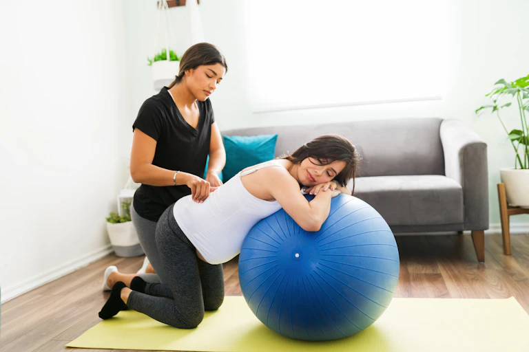 woman massages pregnant woman's back as she leans on yoga ball