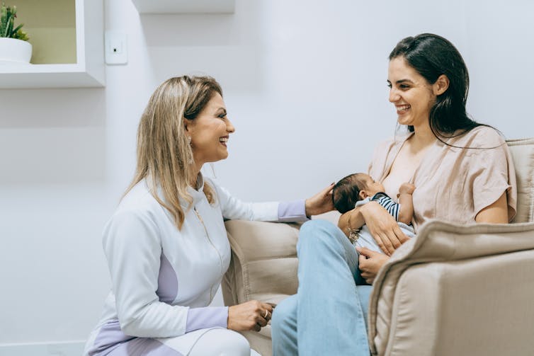 A woman kneels in front of another woman breastfeeding a baby