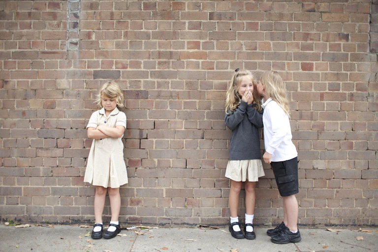 Two children whisper to each other in front of another child who has their arms crossed.