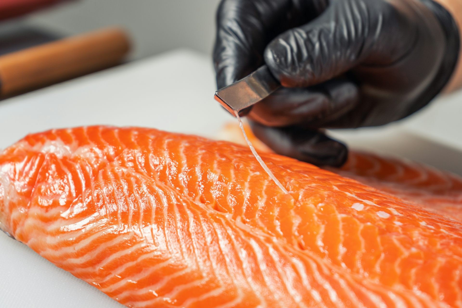 A chef wearing black gloves removes a fish bone from a salmon fillet with tweezers.