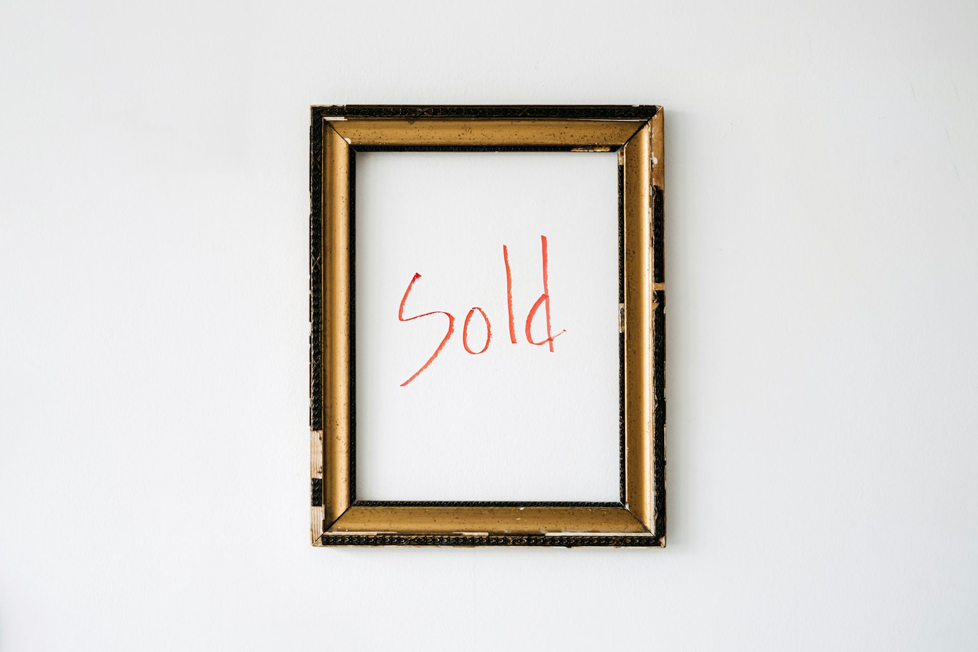 An empty gold-coloured frame hangs on a white wall, with the word "sold" written in red marker within the frame.