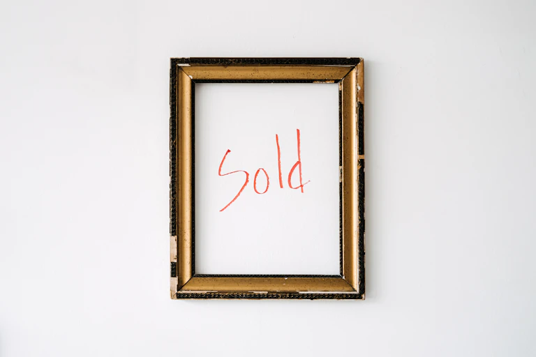 An empty gold-coloured frame hangs on a white wall, with the word "sold" written in red marker within the frame.