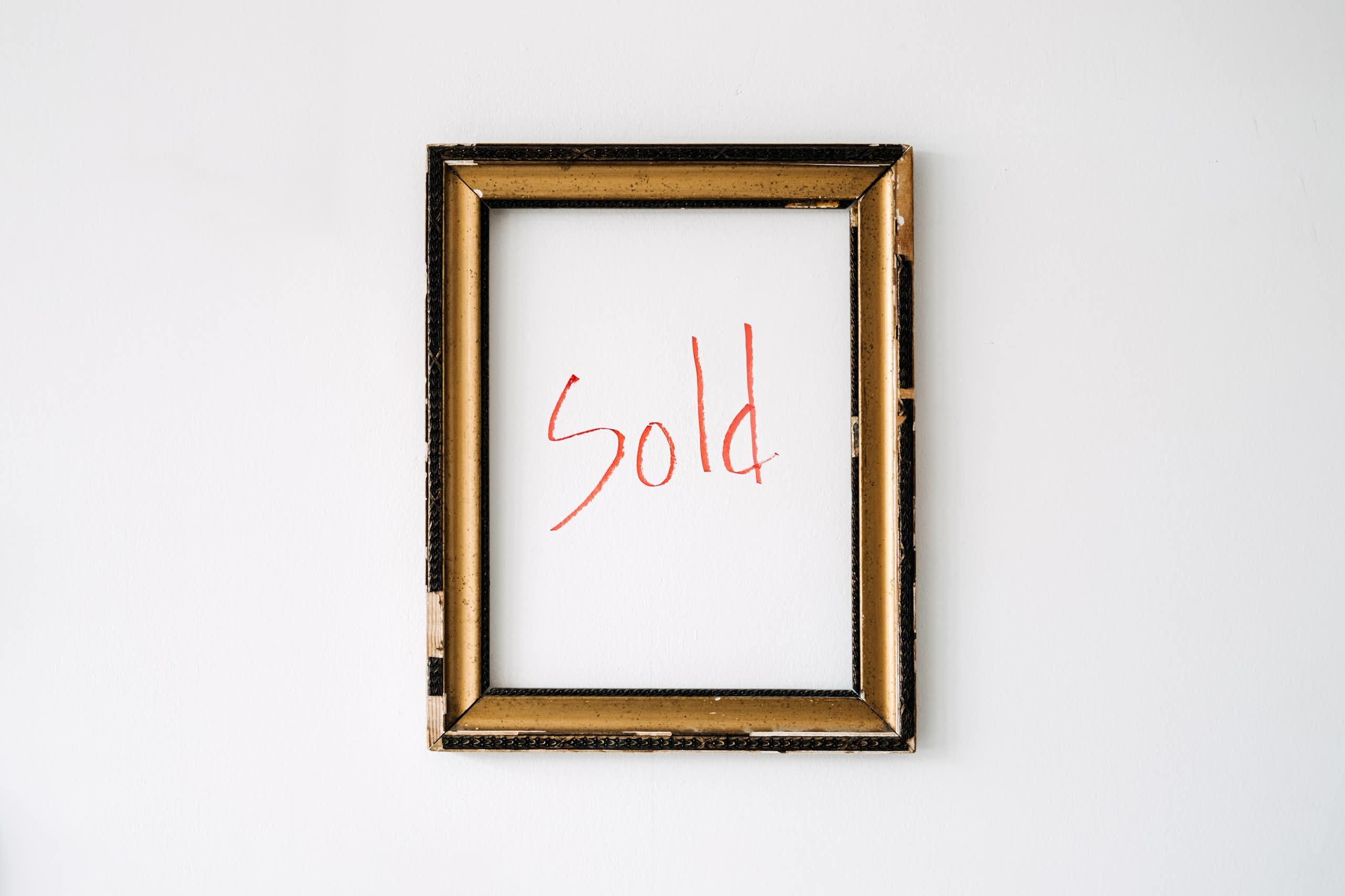 An empty gold-coloured frame hangs on a white wall, with the word "sold" written in red marker within the frame.
