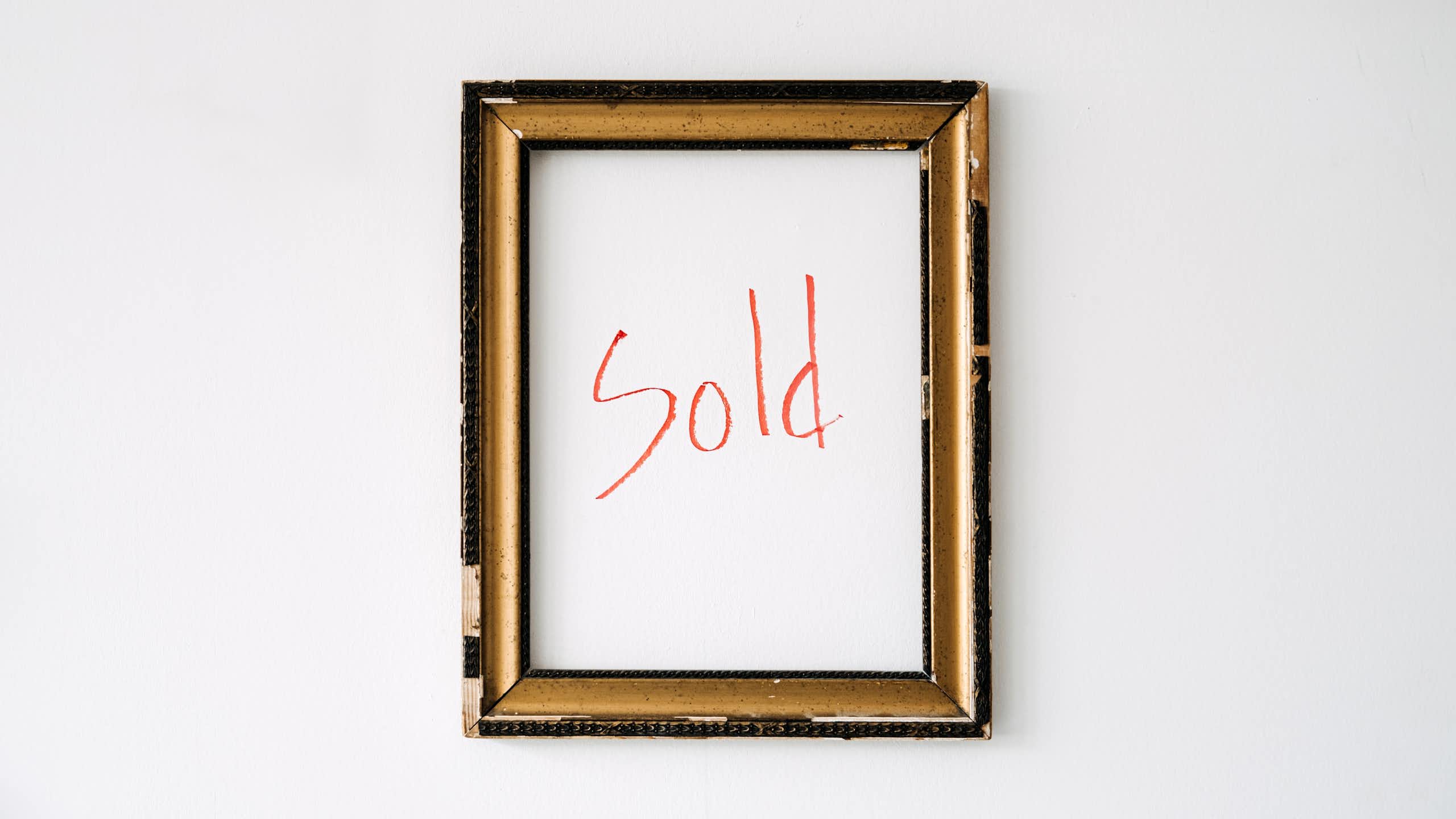 An empty gold-coloured frame hangs on a white wall, with the word "sold" written in red marker within the frame.