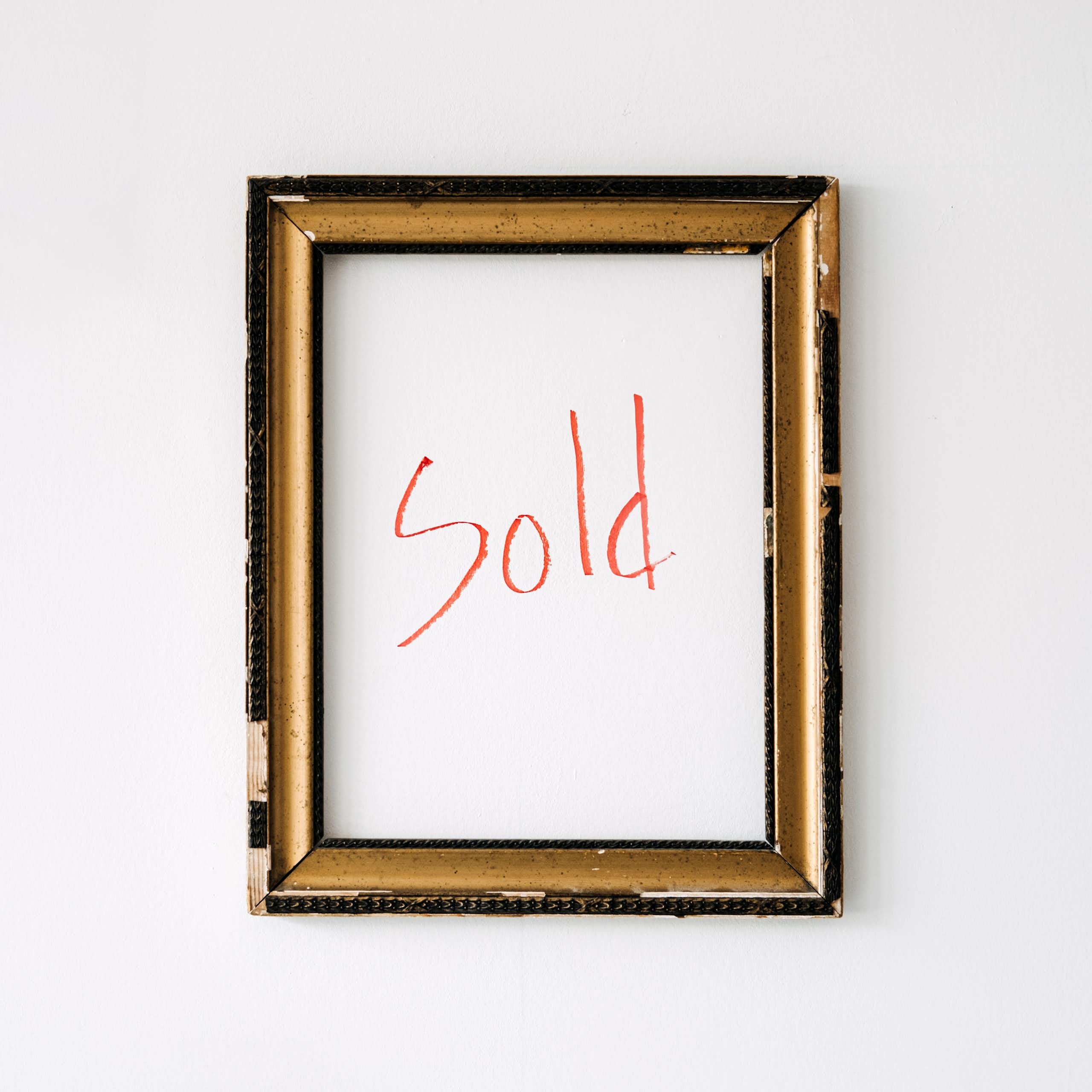 An empty gold-coloured frame hangs on a white wall, with the word "sold" written in red marker within the frame.