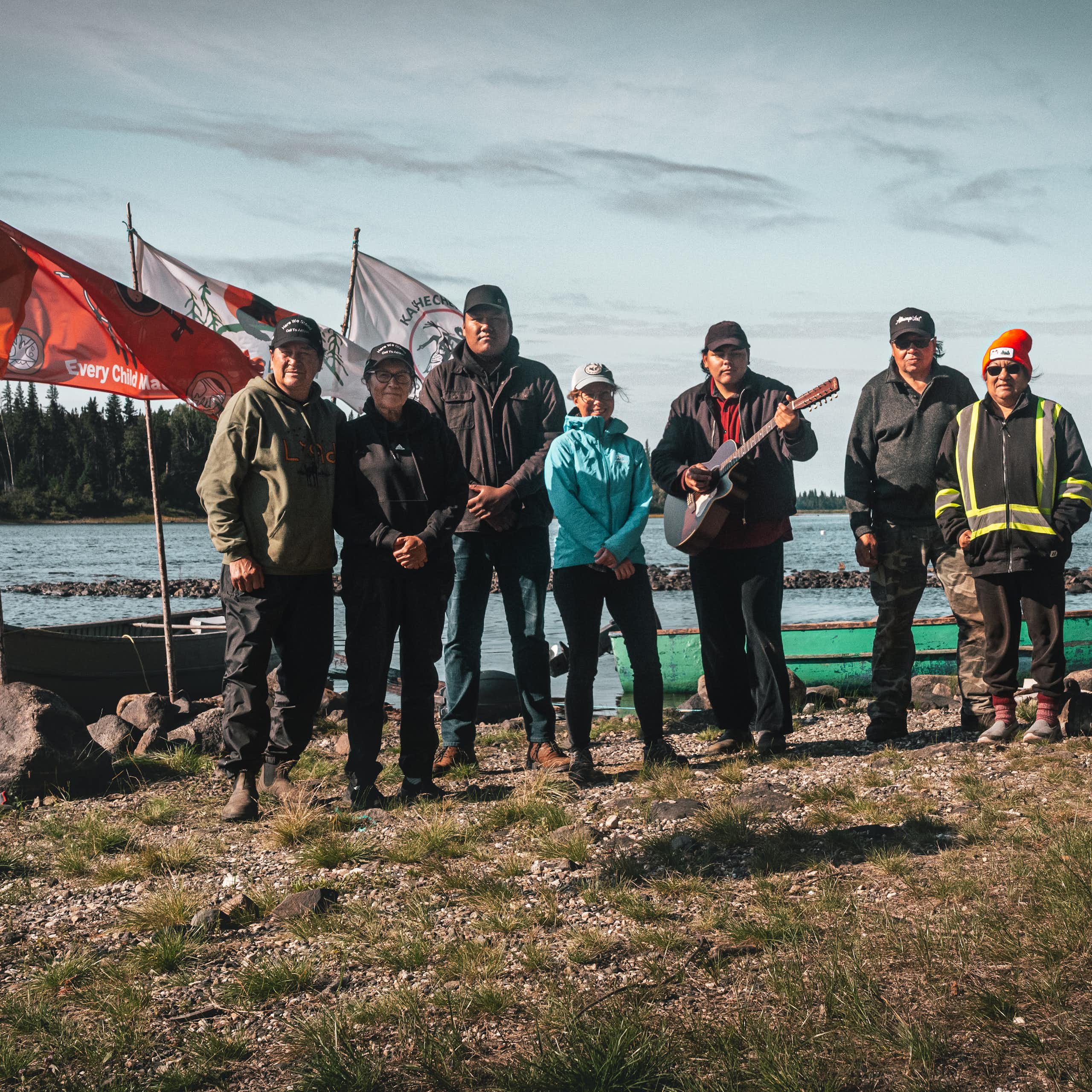 a group of people stand posing for a photo near a body of water. Flag fly of poles behind them
