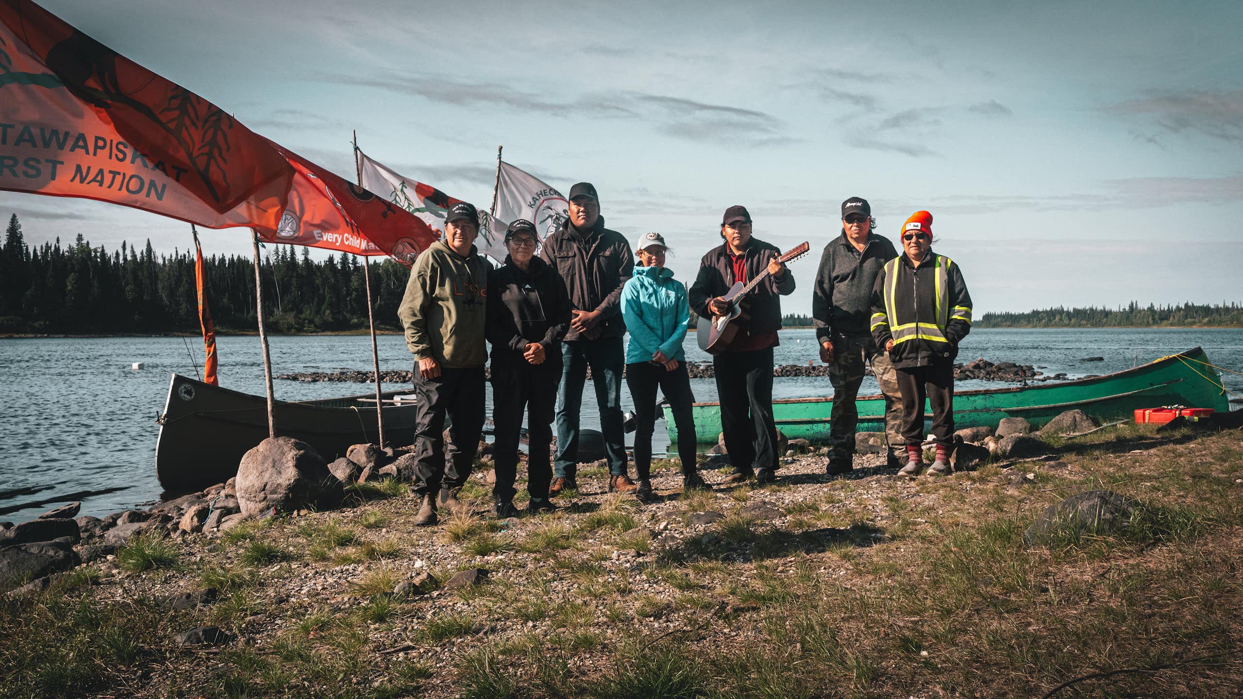 a group of people stand posing for a photo near a body of water. Flag fly of poles behind them