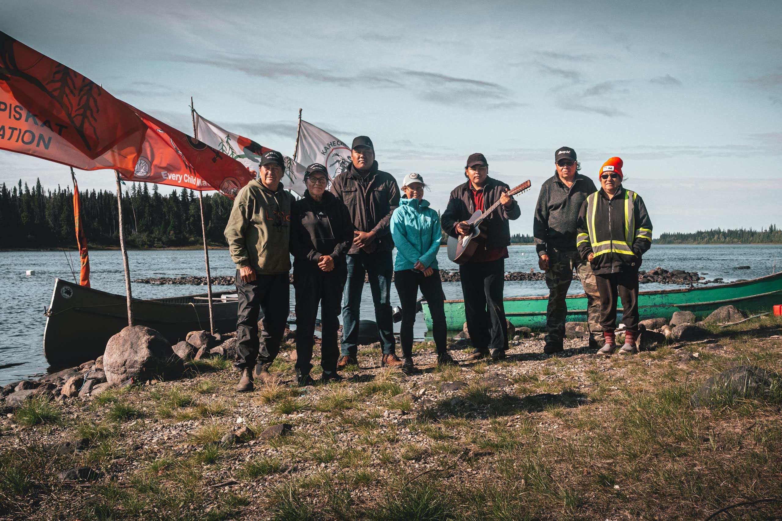 a group of people stand posing for a photo near a body of water. Flag fly of poles behind them