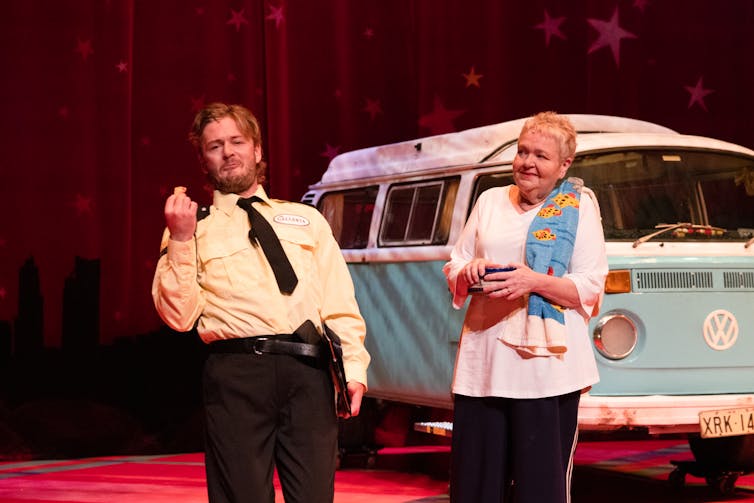 A man in a security uniform stands beside a woman holding a mug, with a towel over her shoulder, in front of a light-blue Volkswagen van.