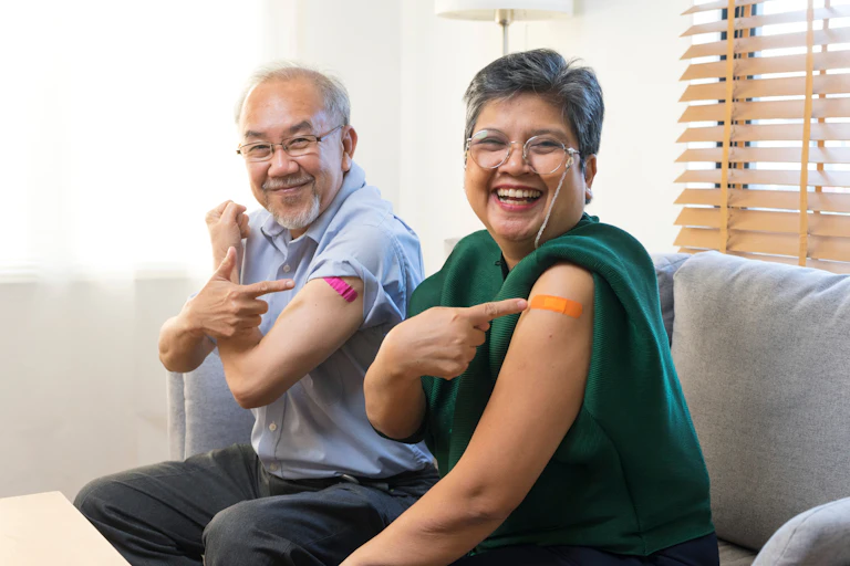 An elderly man and woman pointing at the plasters on their upper arms.