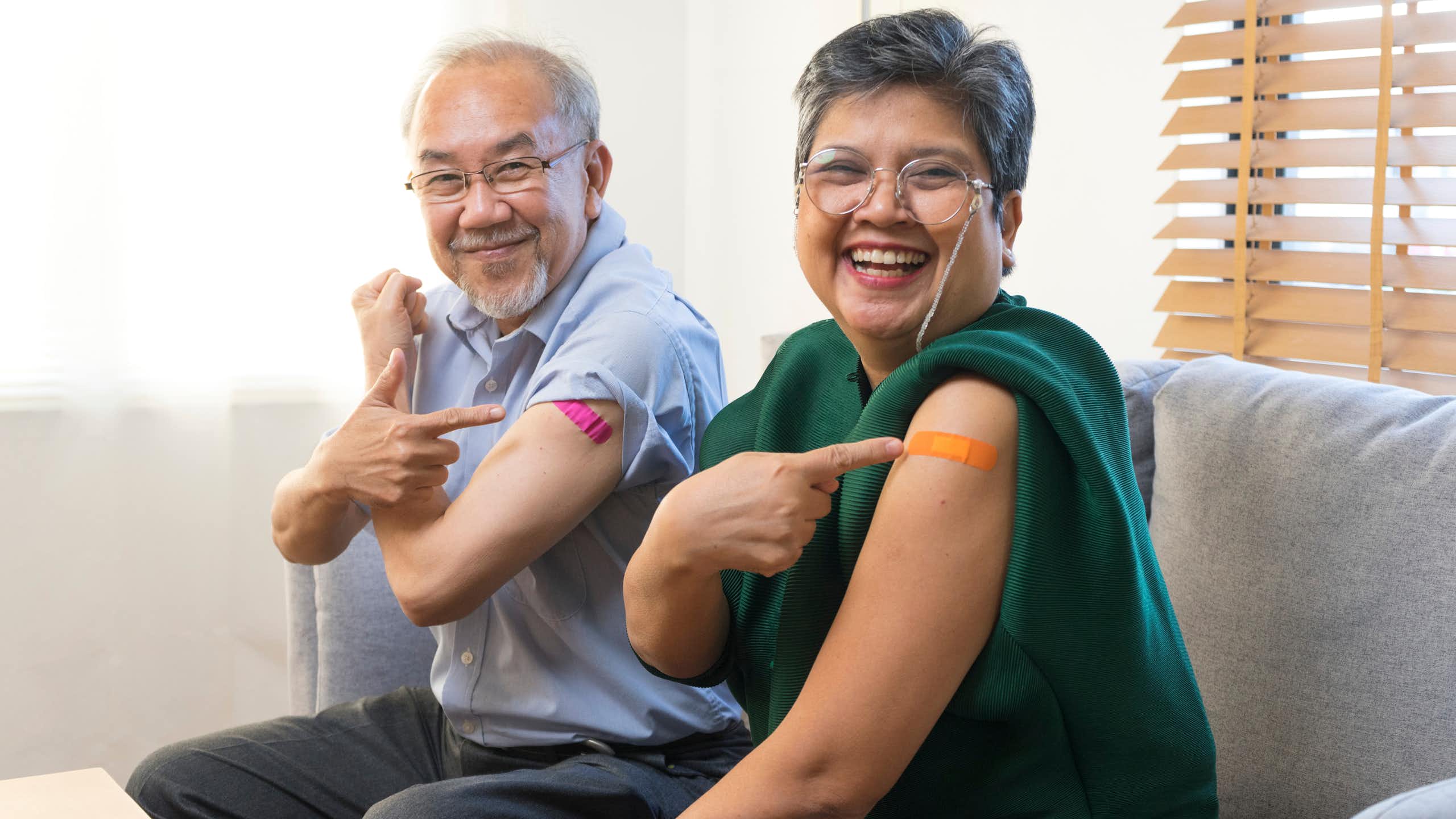 An elderly man and woman pointing at the plasters on their upper arms.
