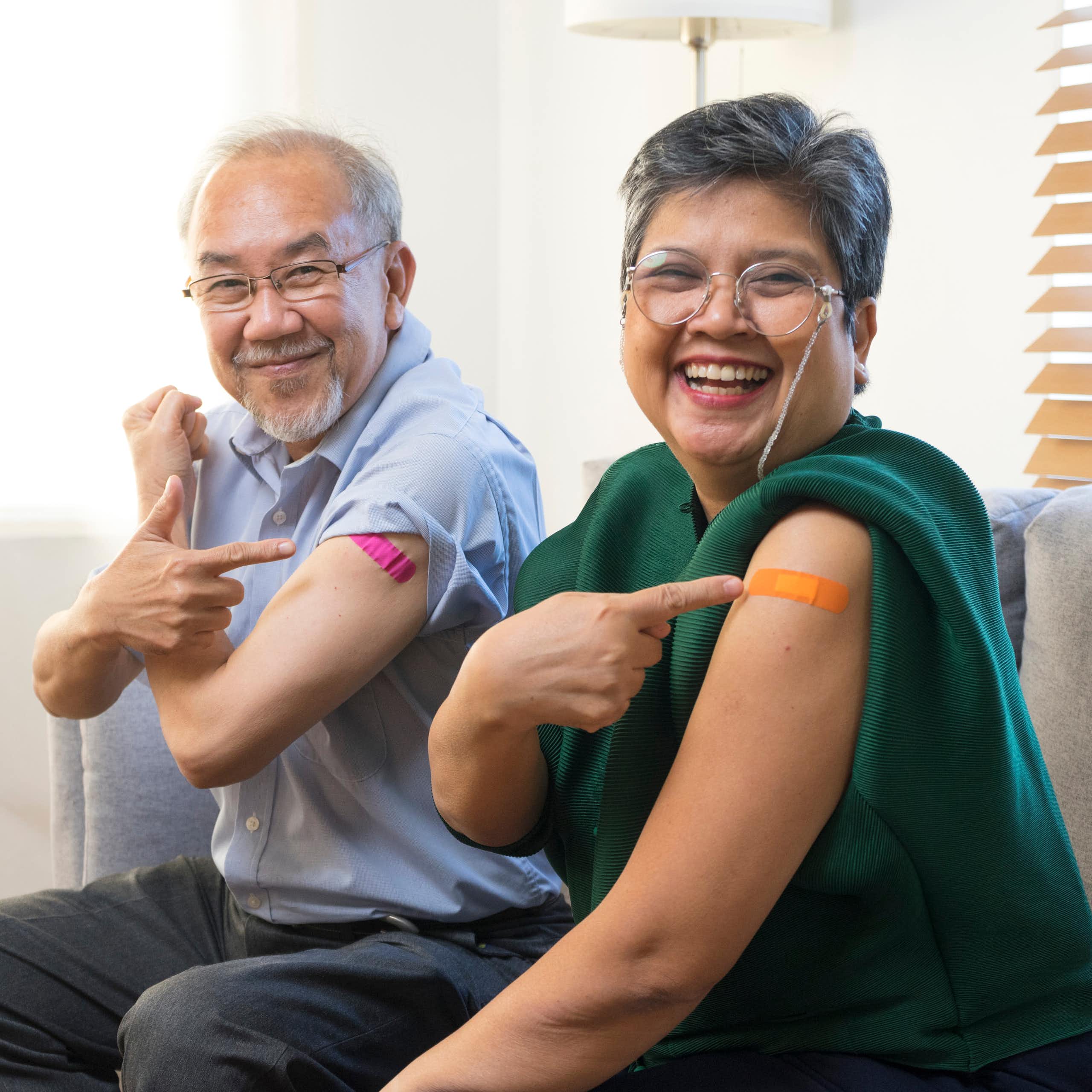 An elderly man and woman pointing at the plasters on their upper arms.