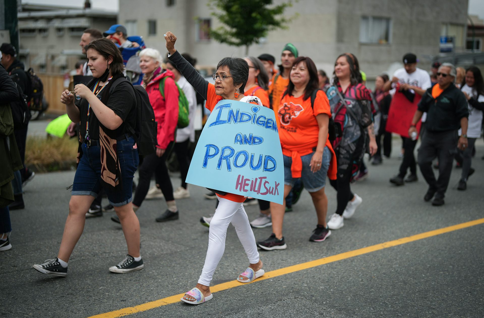 People walk carrying signs and one woman lifts her arm while carrying a sign that says ‘Indigenous and proud.’