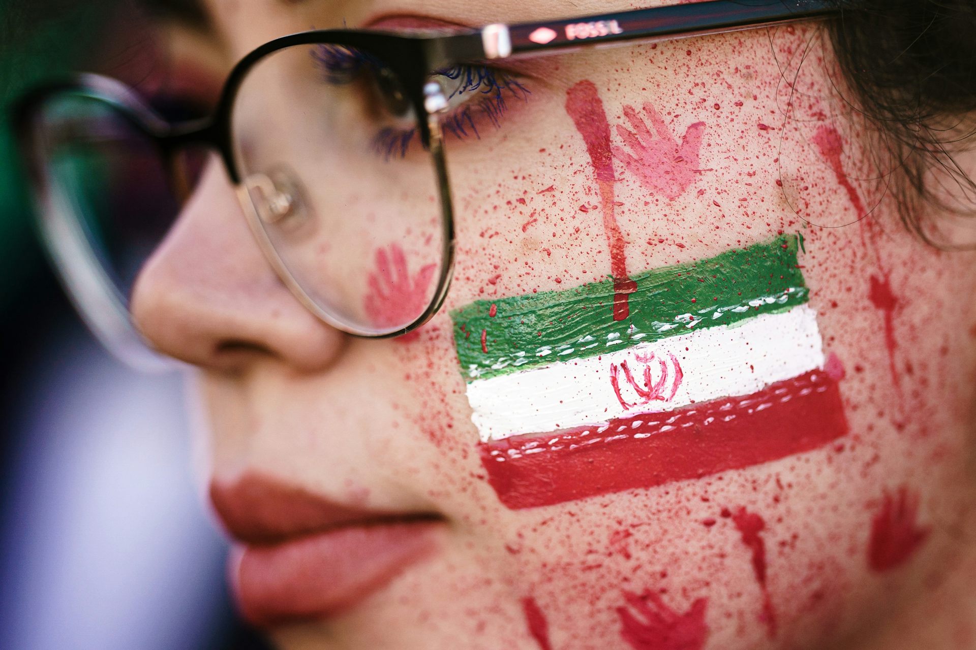A woman with red paint in her face, resembling blood splatters and bloody handprints next to the national Iranian flag.
