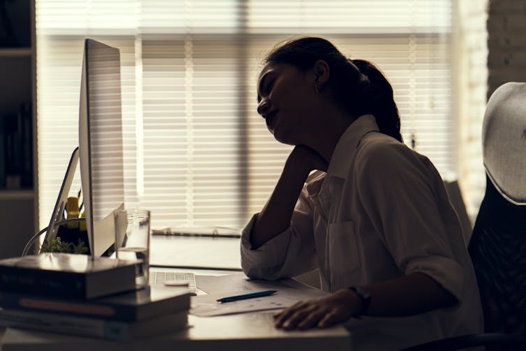 A young woman rubs her neck while sitting in front of a computer in an office