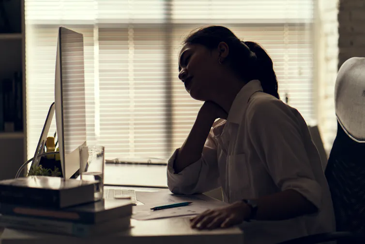 A young woman rubs her neck while sitting in front of a computer in an office