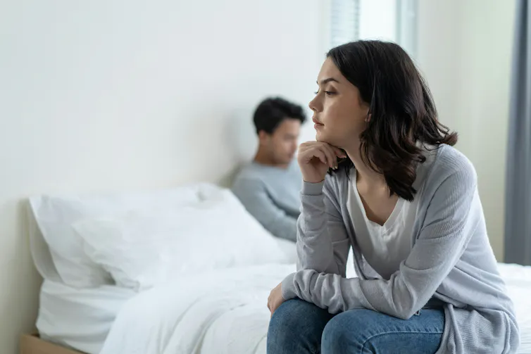 A couple sitting on opposite sides of the bed and looking away from each other.