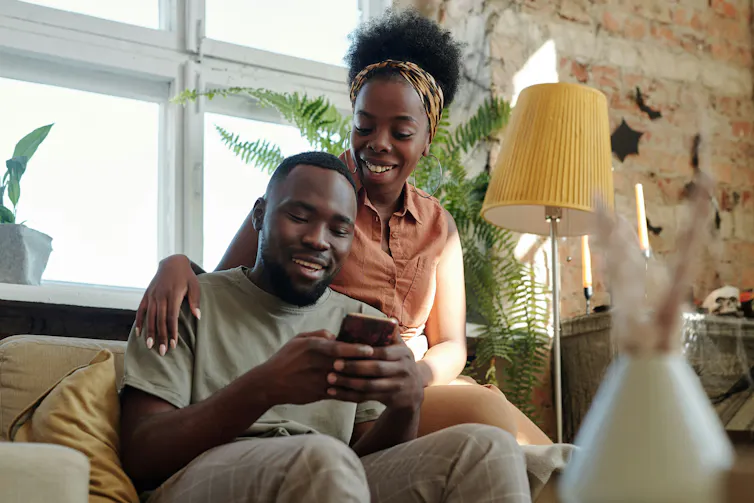 A man sits on a couch using a phone, a woman sits on the arm of the couch behind him, both are smiling.
