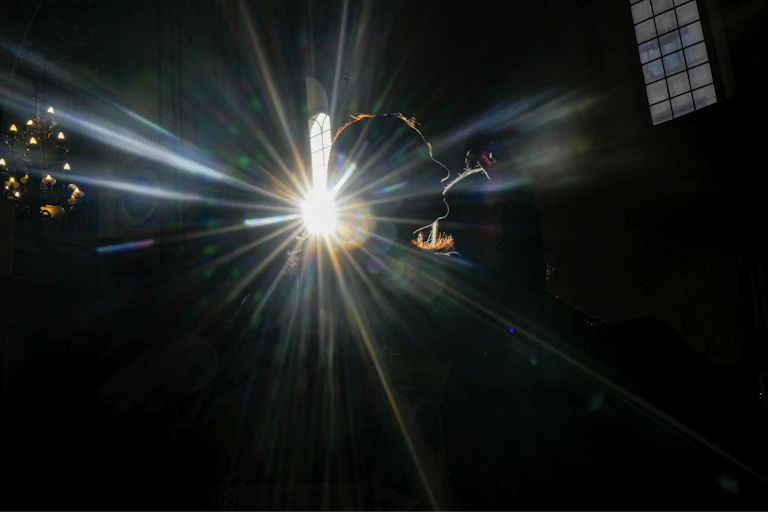 The outline of a woman's face is illuminated in a dark church by a sunbeam coming through a tall window.