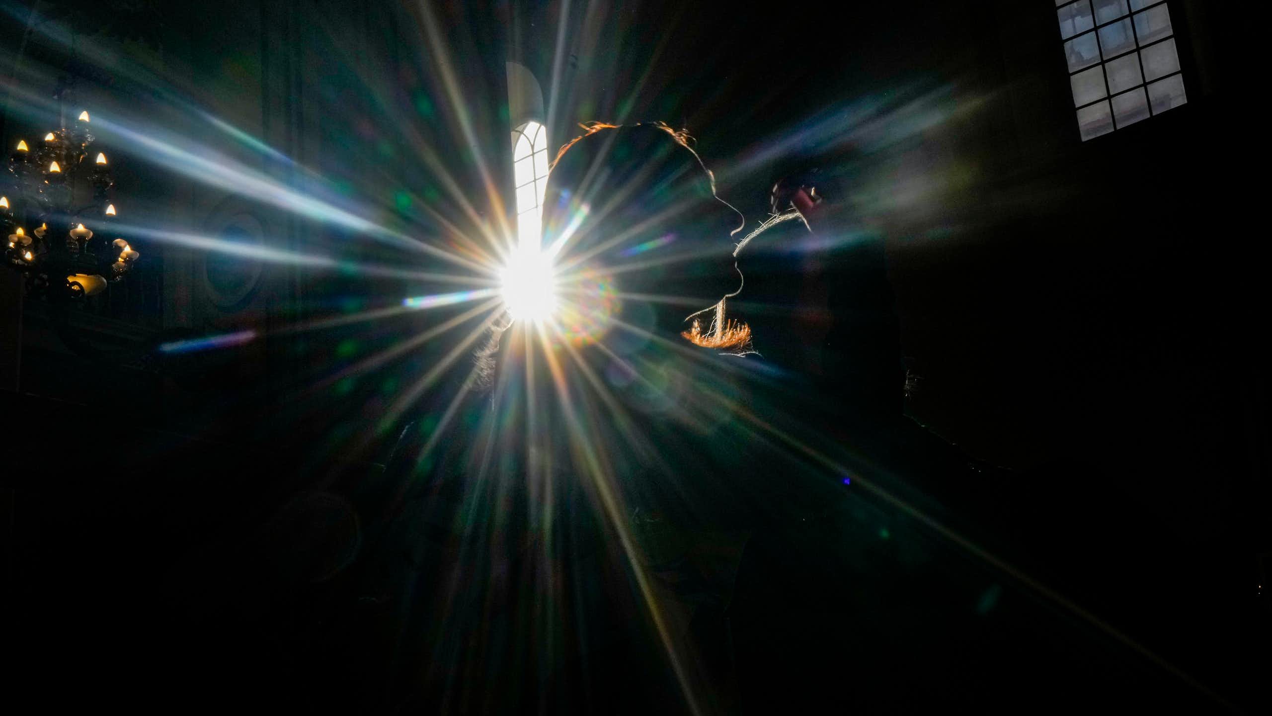 The outline of a woman's face is illuminated in a dark church by a sunbeam coming through a tall window.