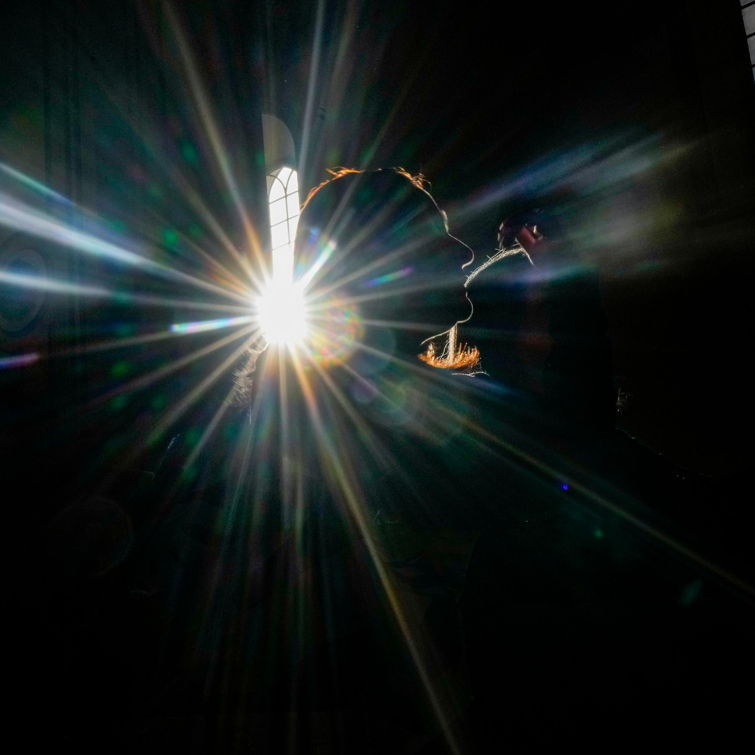 The outline of a woman's face is illuminated in a dark church by a sunbeam coming through a tall window.
