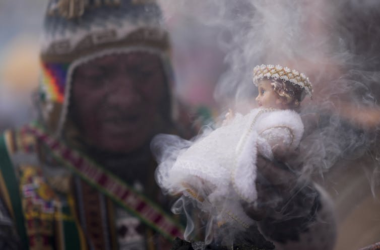 ‘Yes’ to God, however ‘no’ to church – what spiritual alternate seems like for lots of Latin American citizens 2 A man in a colorful knit hat and bright sweater or jacket holds up a small doll in a white robe that is surrounded by wisps of smoke.