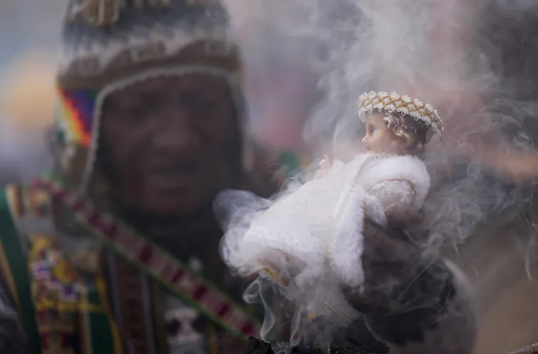 A man in a colorful knit hat and bright sweater or jacket holds up a small doll in a white robe that is surrounded by wisps of smoke.