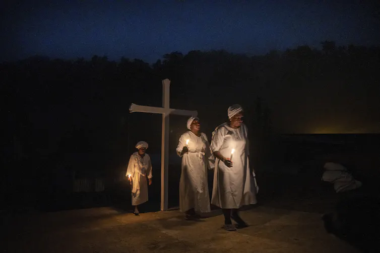 Three women in white robes and caps stand outdoors at nighttime by a large wooden cross.