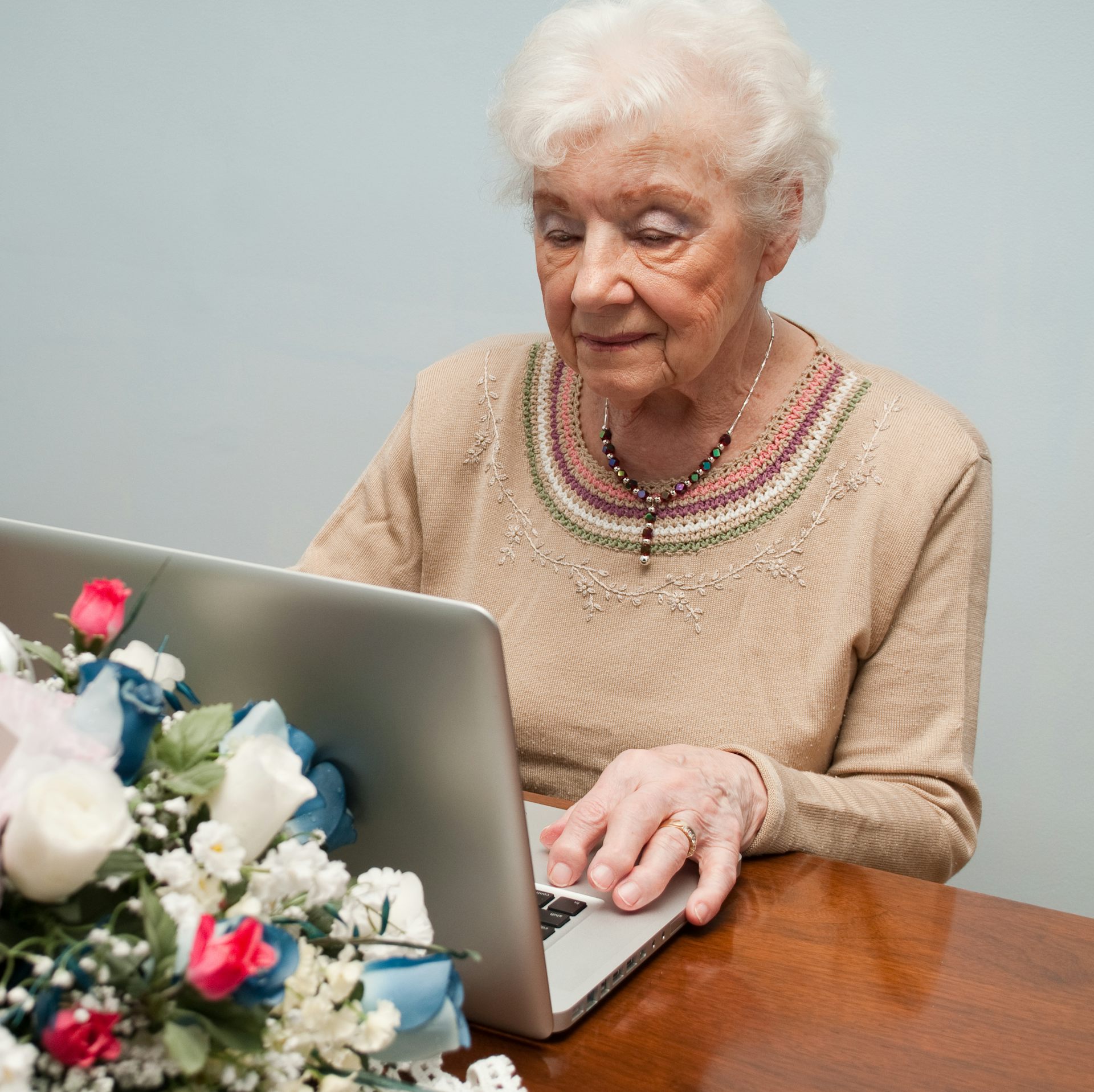 An elderly woman works on a laptop, looking grim, with a bouquet of flowers behind it.