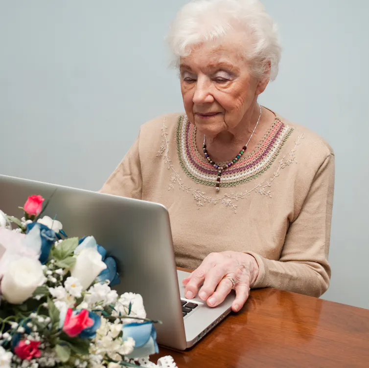 An elderly woman works on a laptop, looking grim, with a bouquet of flowers behind it.
