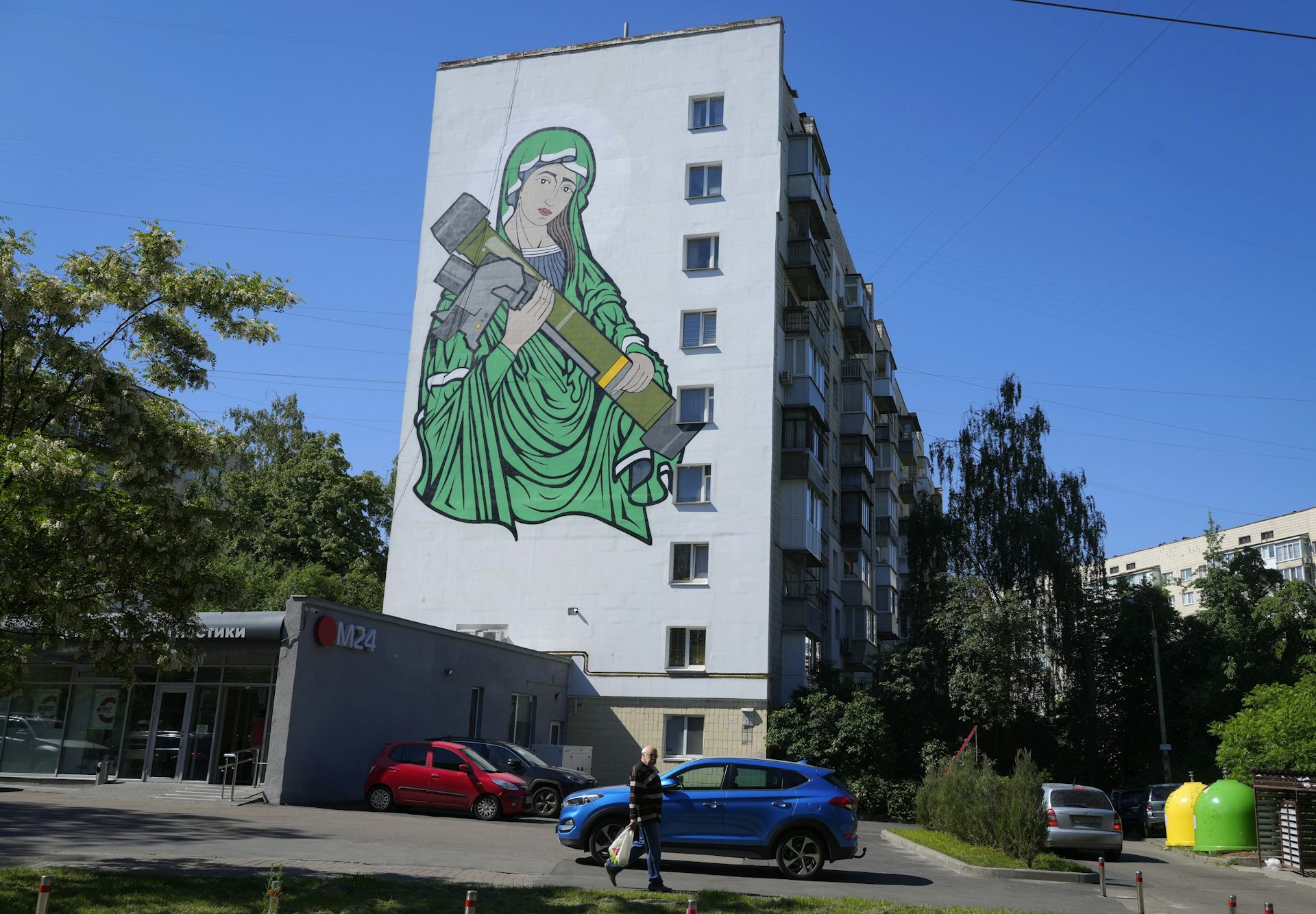 A painting of a woman in green robes cradling an anti-tank weapon on the side of an apartment building.