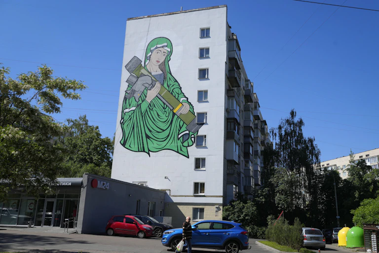 A painting of a woman in green robes cradling an anti-tank weapon on the side of an apartment building.