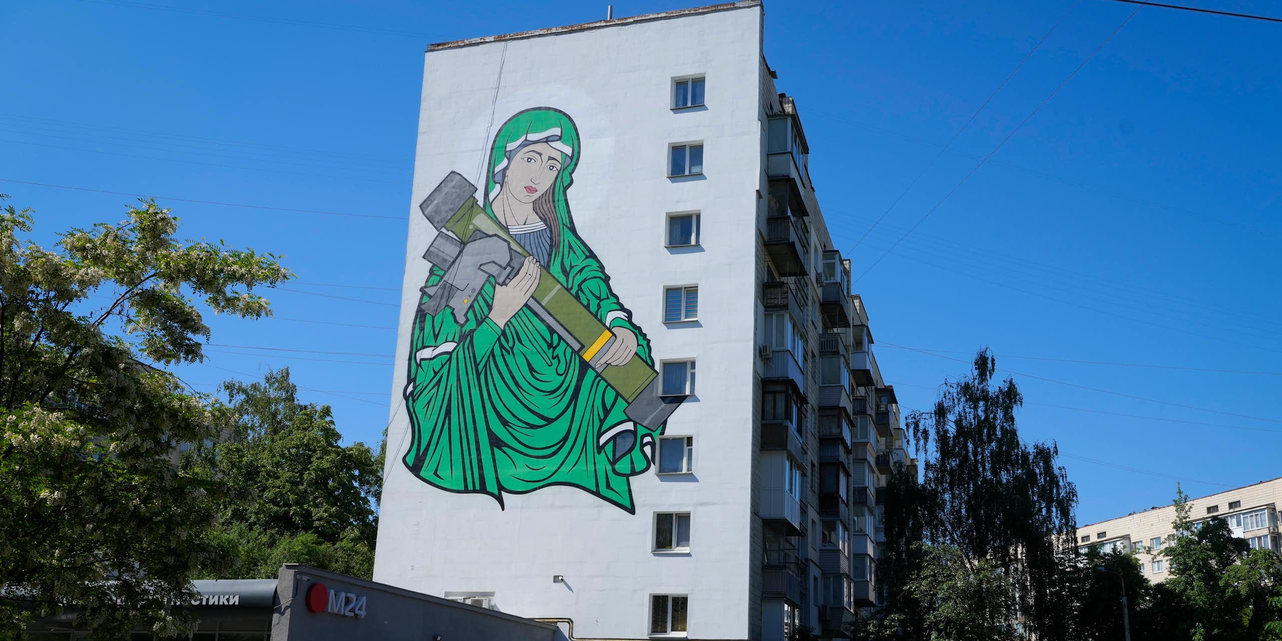A painting of a woman in green robes cradling an anti-tank weapon on the side of an apartment building.