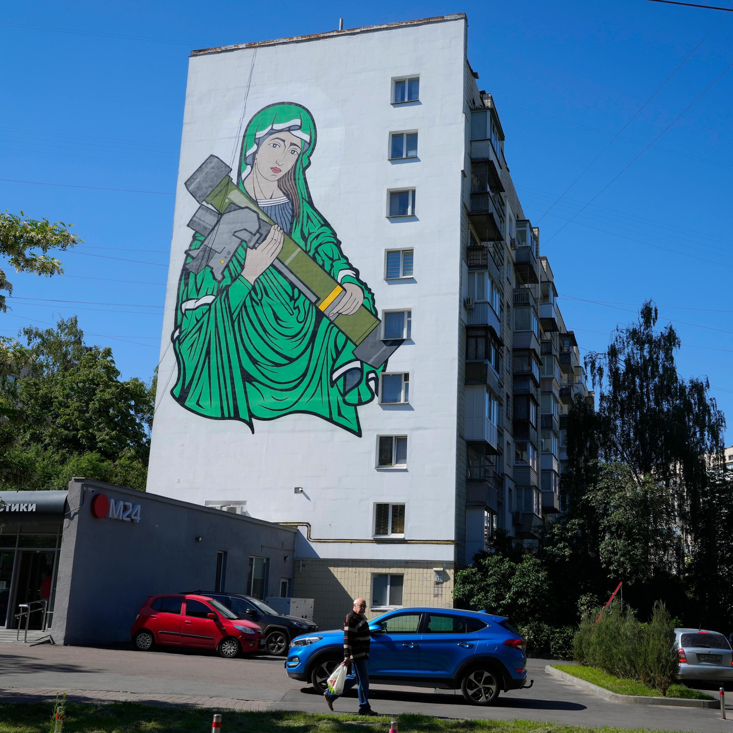 A painting of a woman in green robes cradling an anti-tank weapon on the side of an apartment building.