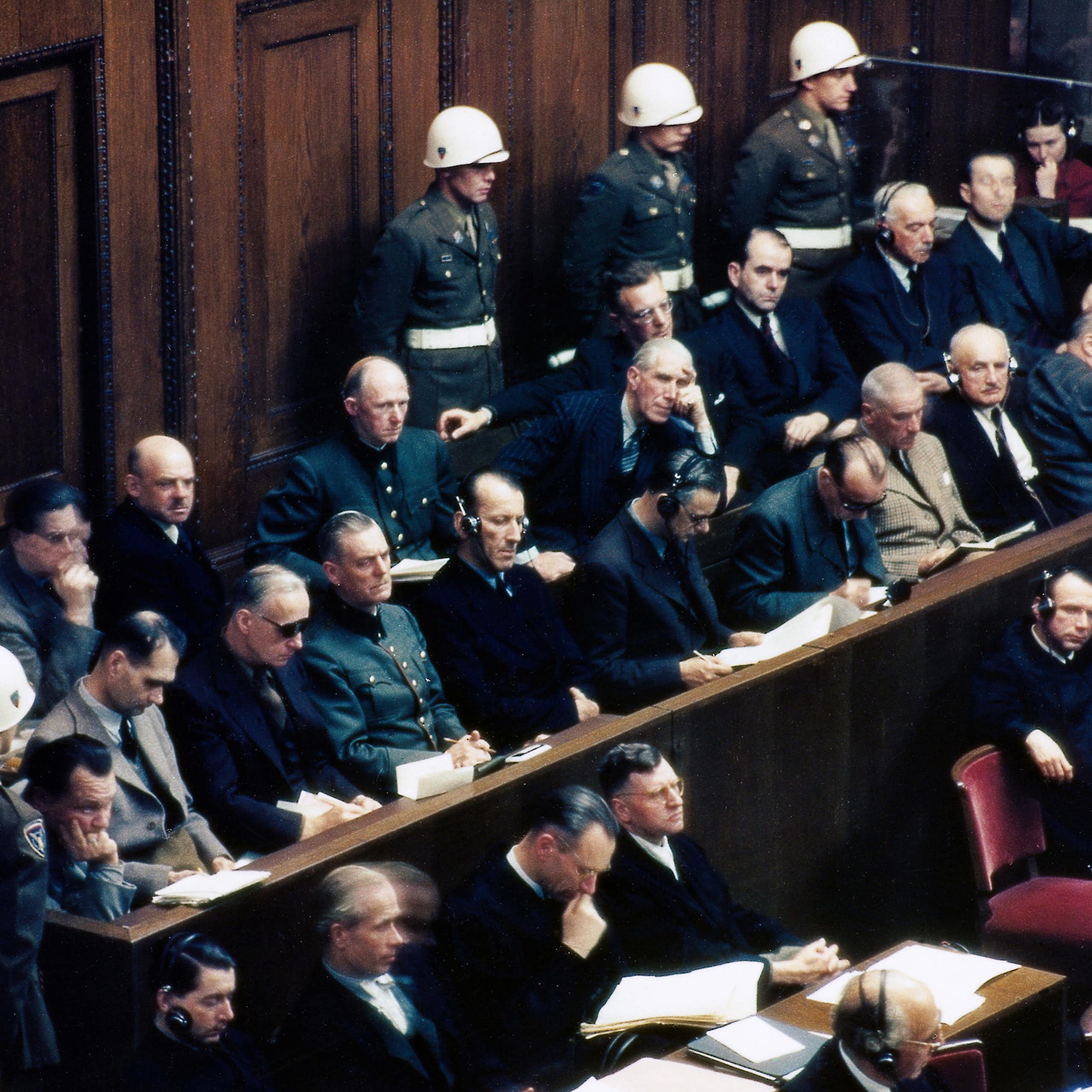 A courtroom with military police standing against a back wall behind two rows of seated defendants.