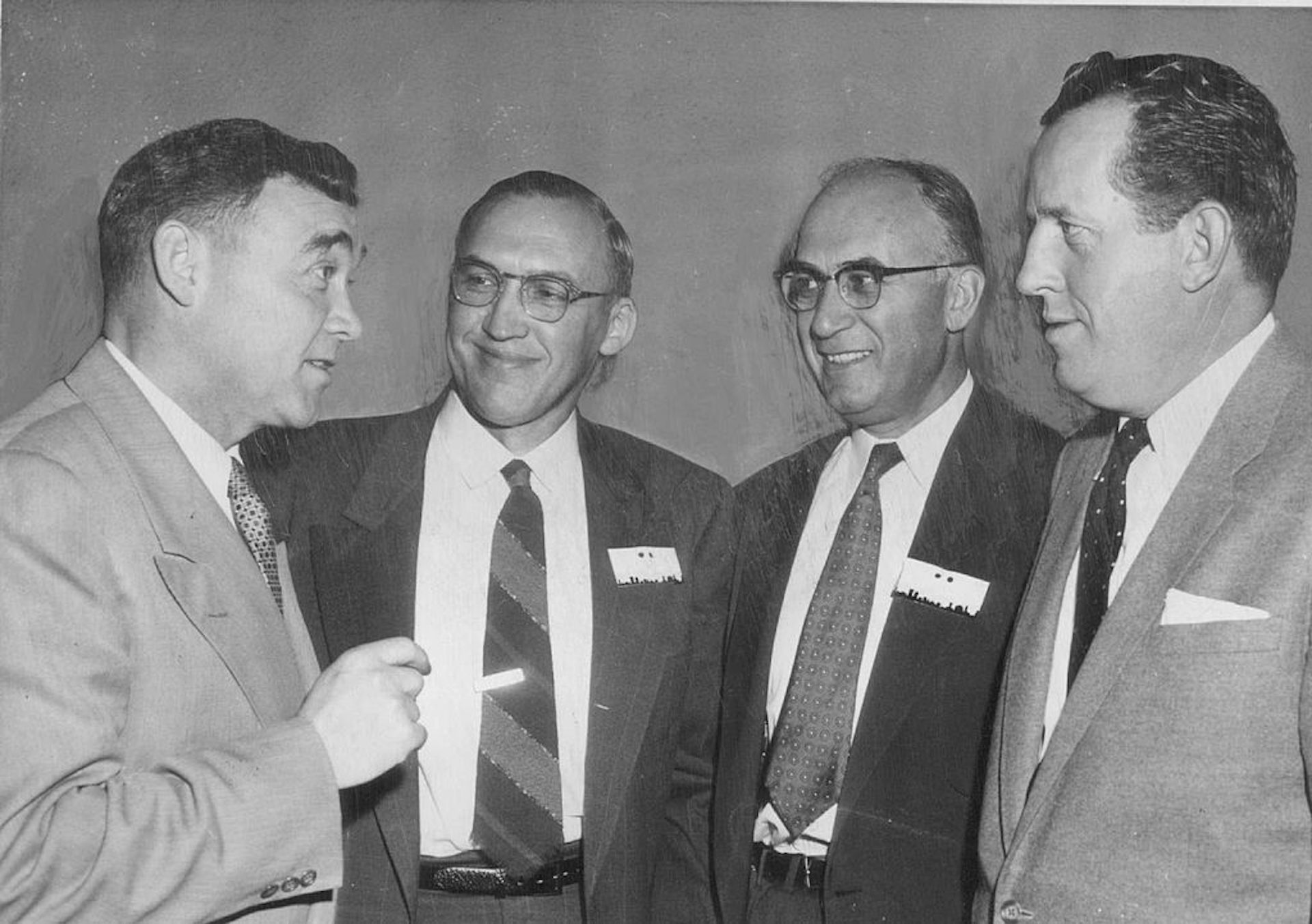 Black-and-white photo of four middle-aged men chatting while wearing suits and ties.