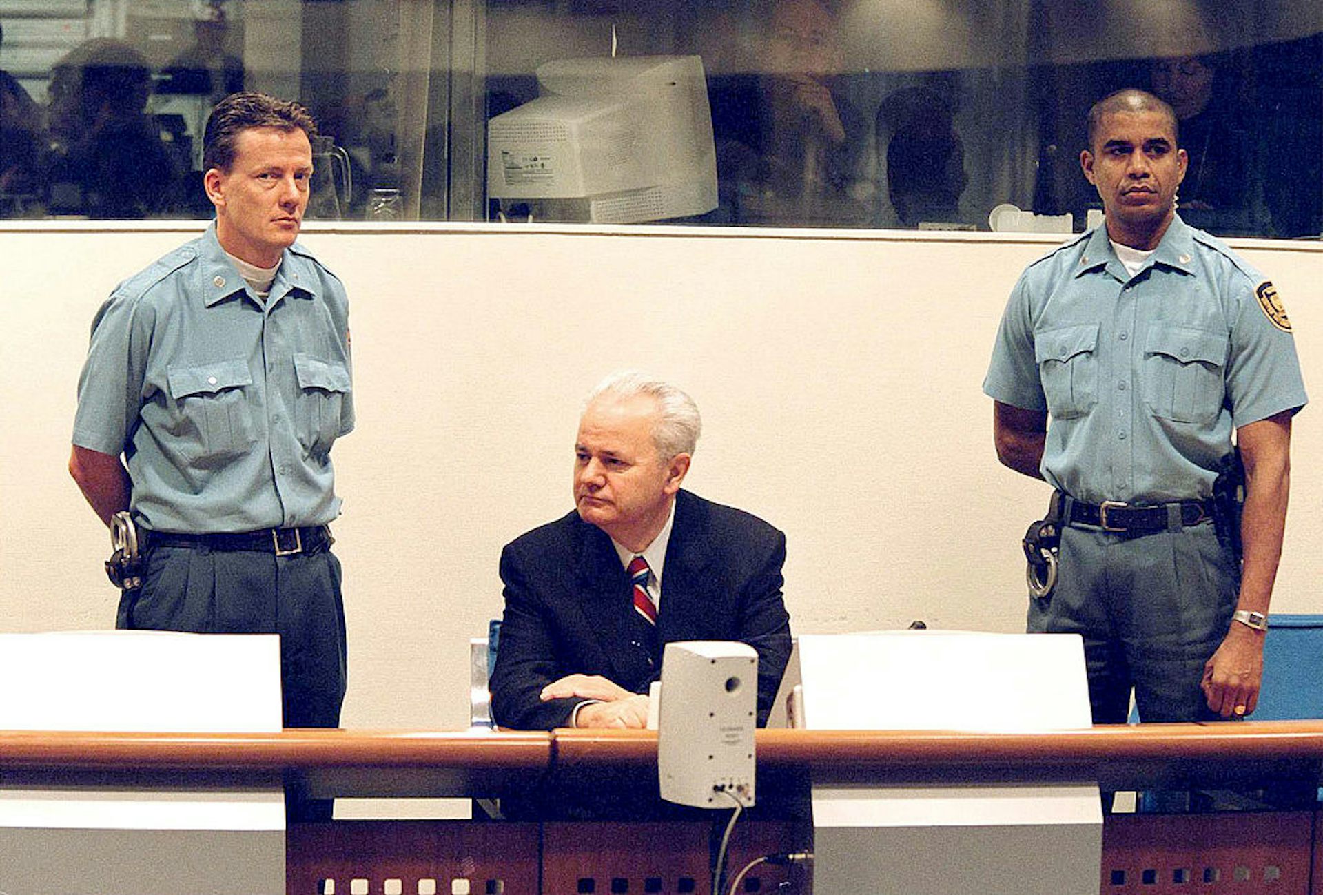 Man with white hair wearing a suit seated and flanked by two court officers.