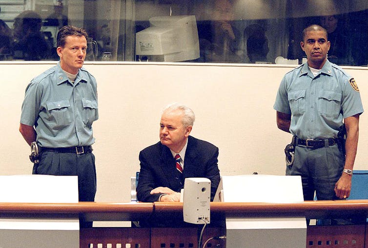 Man with white hair wearing a suit seated and flanked by two court officers.