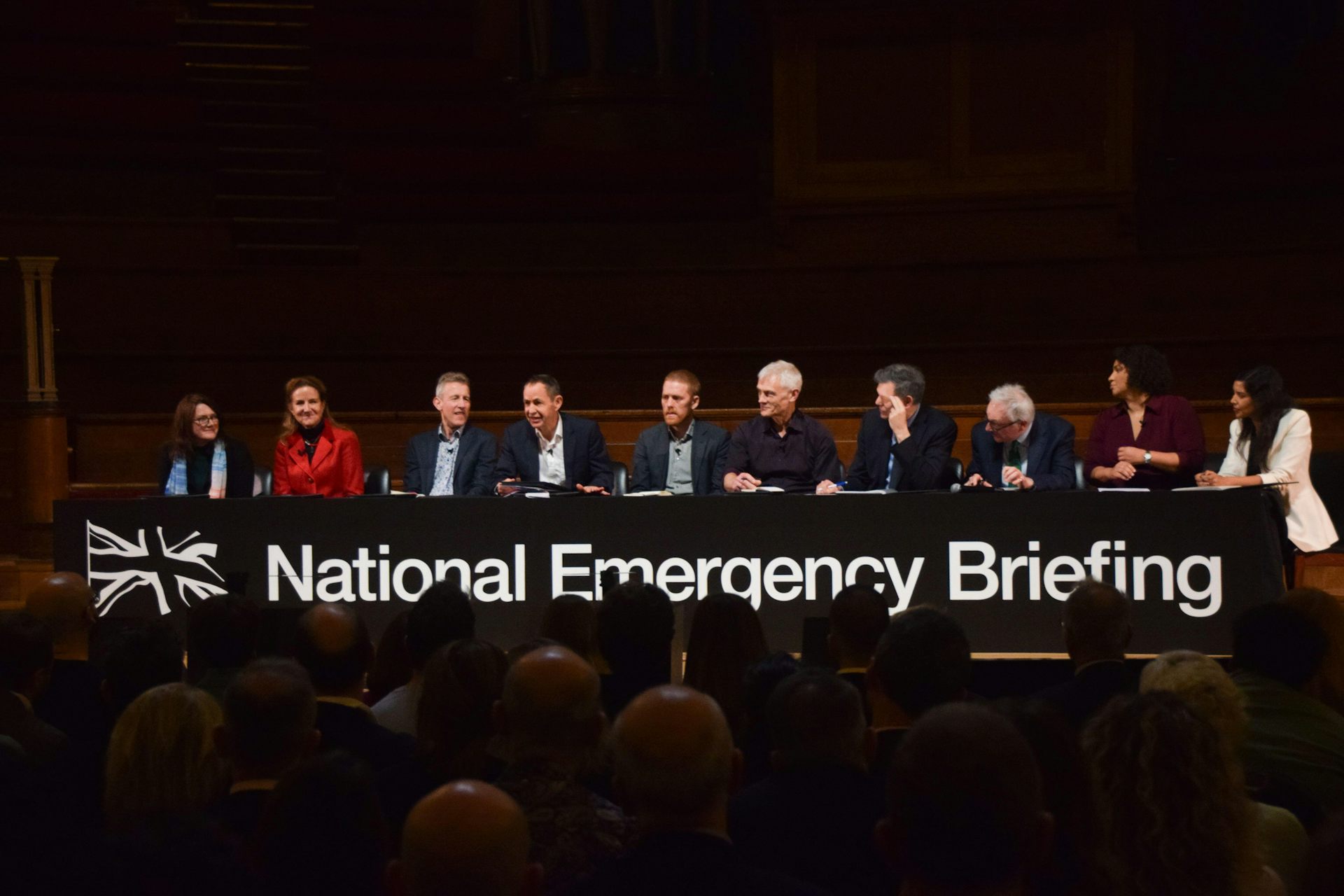line of climate scientist speakers sat at long table, black background, sign says national emergency briefing below them