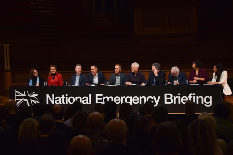 line of climate scientist speakers sat at long table, black background, sign says national emergency briefing below them