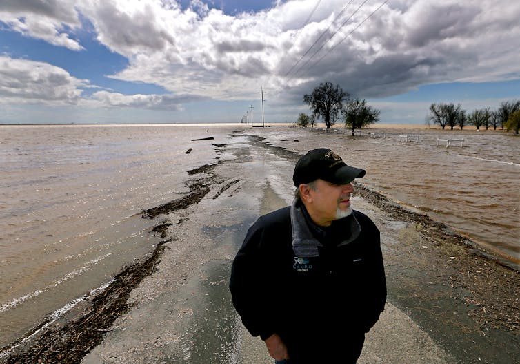 a man stands on a road that is flooded on both sides as far as the camera can see. Trees are surrounded by flood water on one side.