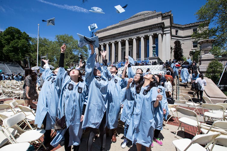 Federal investment cuts are just one drawback going through The usa’s faculties and universities 1 A group of young people wear light blue graduation robes and throw their caps into the air. They stand outside of a large building with columns.