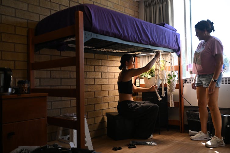 A young woman hangs a plant under a lofted bed with a brick wall against it. Another woman stands nearby her near a window.