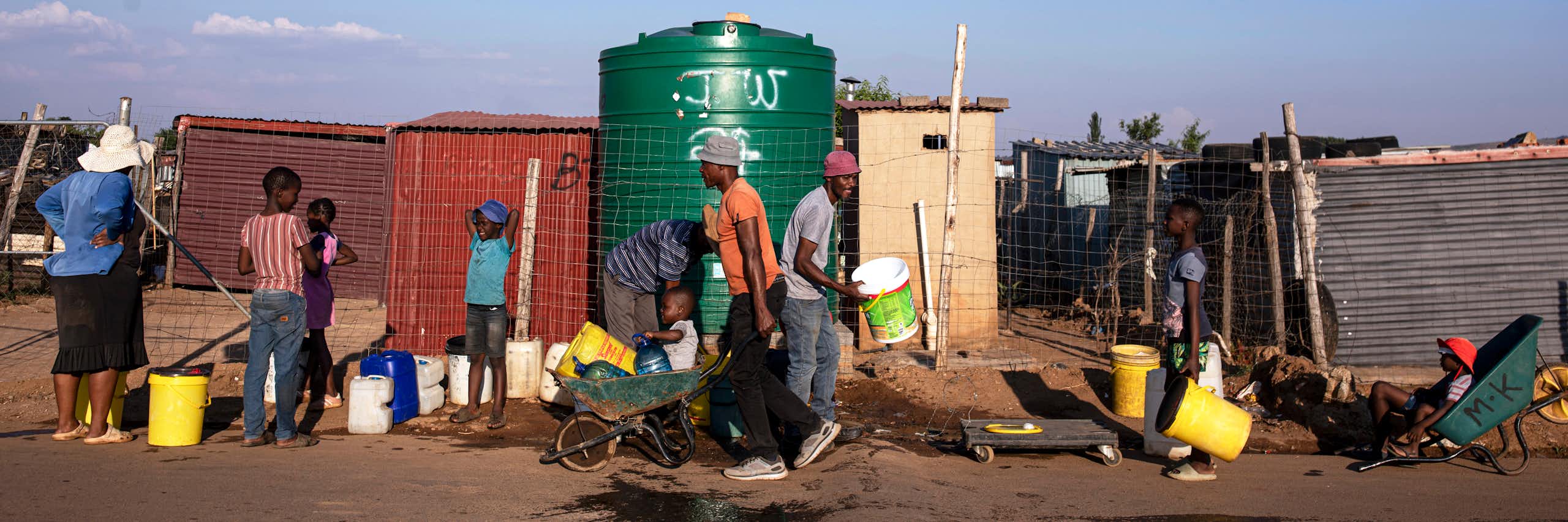 People lining up with buckets and carrying buckets of water in wheelbarrows - lining up at a 5000 litre water tank