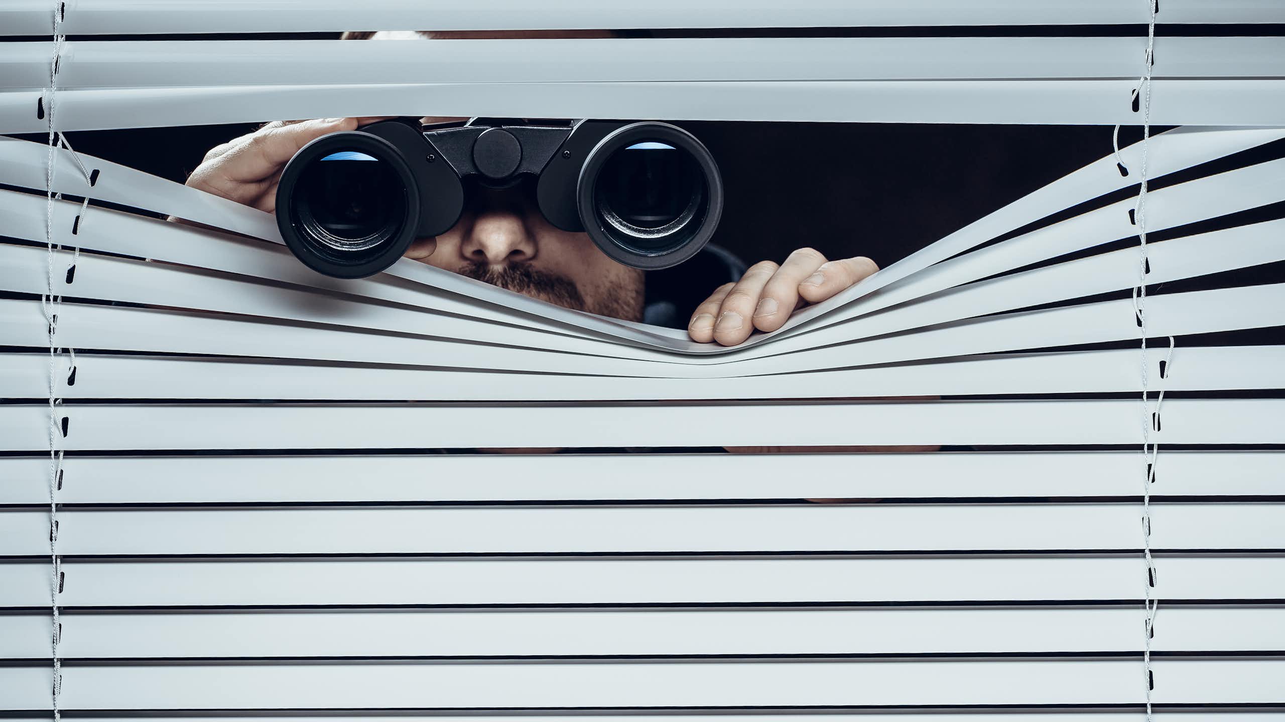 A person using binoculars to secretly look out through a window with blinds in it.